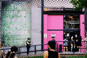 The exterior of Buddies in Bad Times Theatre. The bubblegum-pink double doors are open, allowing a crowd of visitors to enter. Above the door is the company's logo, also in pink. To the left of the door, a large green banner hangs against a concrete wall. Pale pink text on the banner reads "THE FUTURE IS RISKY AND POSSIBLE." Image courtesy of the Buddies in Bad Times website.