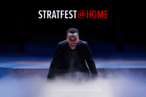 Amaka Umeh as the titular prince in Stratford Festival's 2022 production of Hamlet. She crouches onstage, facing the camera, shouting intently at her audience. Smoke billows from the stage around her hands. Above her head is the red and white logo for STRATFEST@HOME. Original Photo by David Hou. Image courtesy of Stratford Festival.