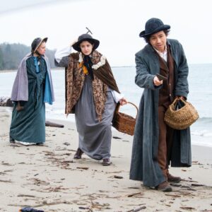 Three women in period clothing walk in on the beach