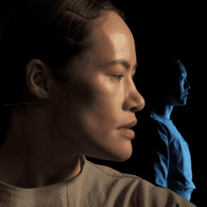 Photo of actor Donna Soares in profile with dramatic lighting. Behind them is another actor cloaked in blue light.
