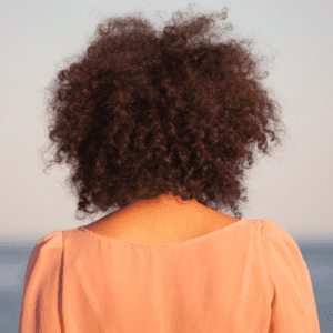 Photo of a woman from behind with tight curly hair. She is overlooking water and her hair is visibly windblown.