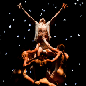 A group of dancers lifting up one dancer with their arms extended up. The background appears like a dark sky with stars.