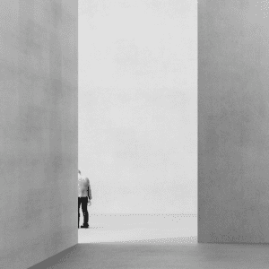 A greyscale image of two large walls with an opening in the middle. The form a person is visible peaking out from the edge of the left wall.