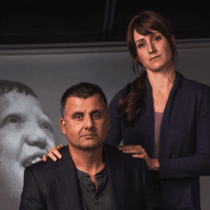 Actor Marcus Youssef sitting with actor Meghan Gardiner standing behind him with her hands on his shoulder. In the background is a projected image of their child in the play.