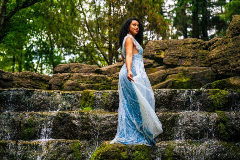 actor vanessa sears stands on a waterfall in a sparkly blue evening dress.