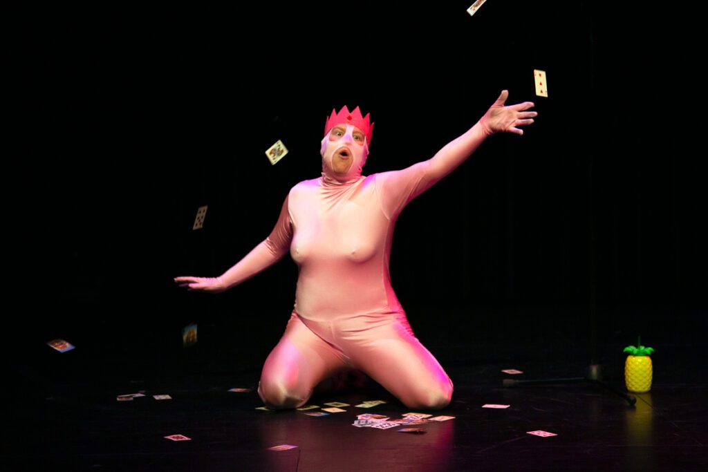 The King of the Party, played by Lou Campbell, kneels on stage
with arms dramatically outstretched. Lou is dressed in a light pink bodysuit and pink-red
crown, with their mouth open as if mid-speech or song. Playing cards are scattered
around and suspended mid-air, adding a sense of motion. A small yellow
pineapple-shaped cup sits on the stage near a microphone stand. The dark background
contrasts sharply with the brightly lit costume and props.